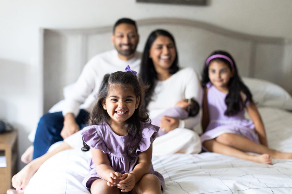Toddler girl sitting on edge of bed in front of mom, dad, older sister and newborn sister in atlanta